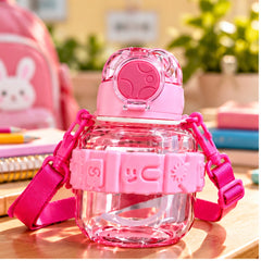 Botella infantil transparente con tapa rosa, correa ajustable y boquilla abatible, ambientada en escritorio