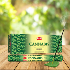 Incienso HEM Cannabis Hexa encendido en soporte de hoja, junto a la caja y las varillas.